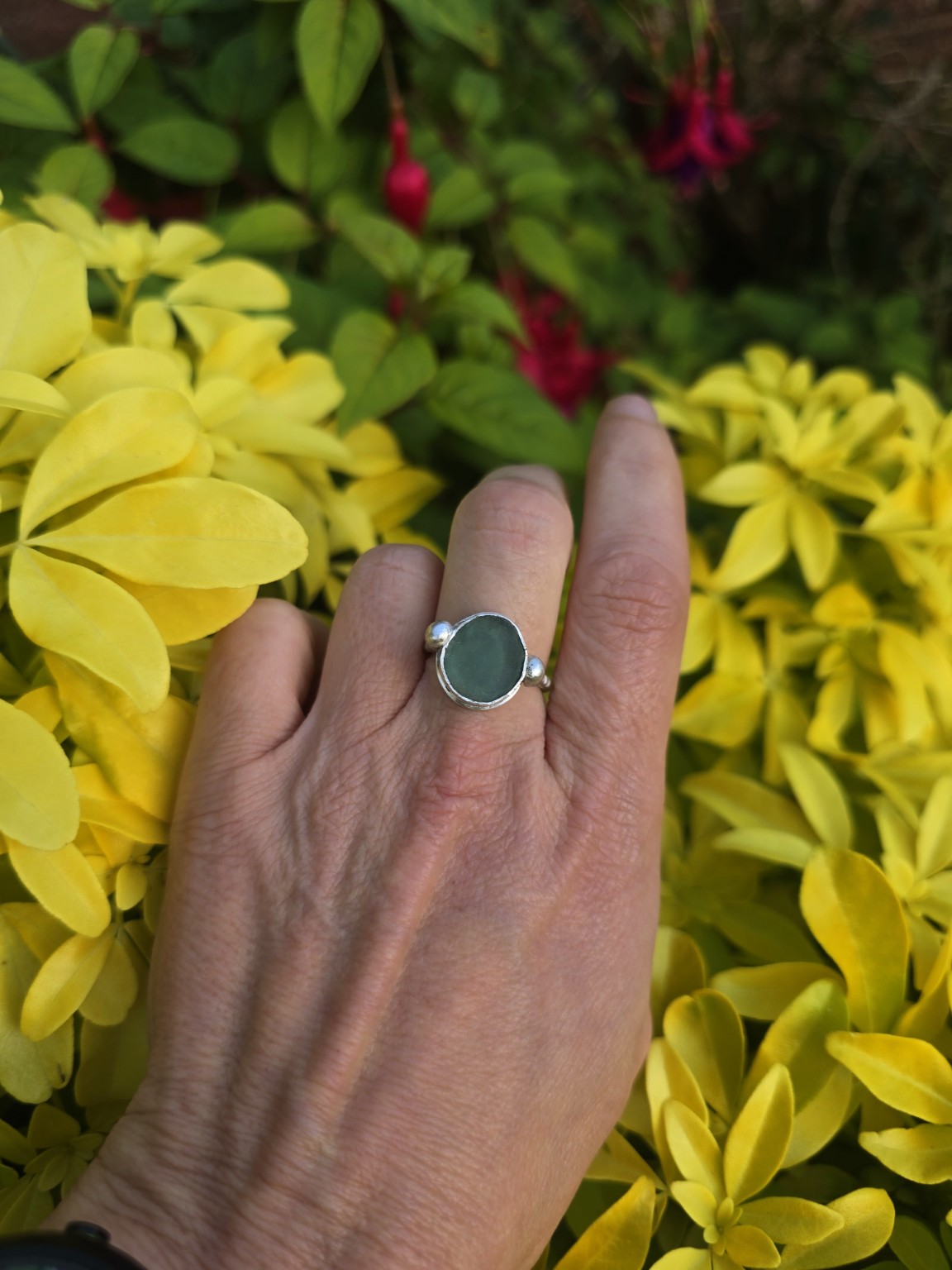 Ocean-blue sea glass ring with silver bobbles on either side, worn on a hand against yellow foliage